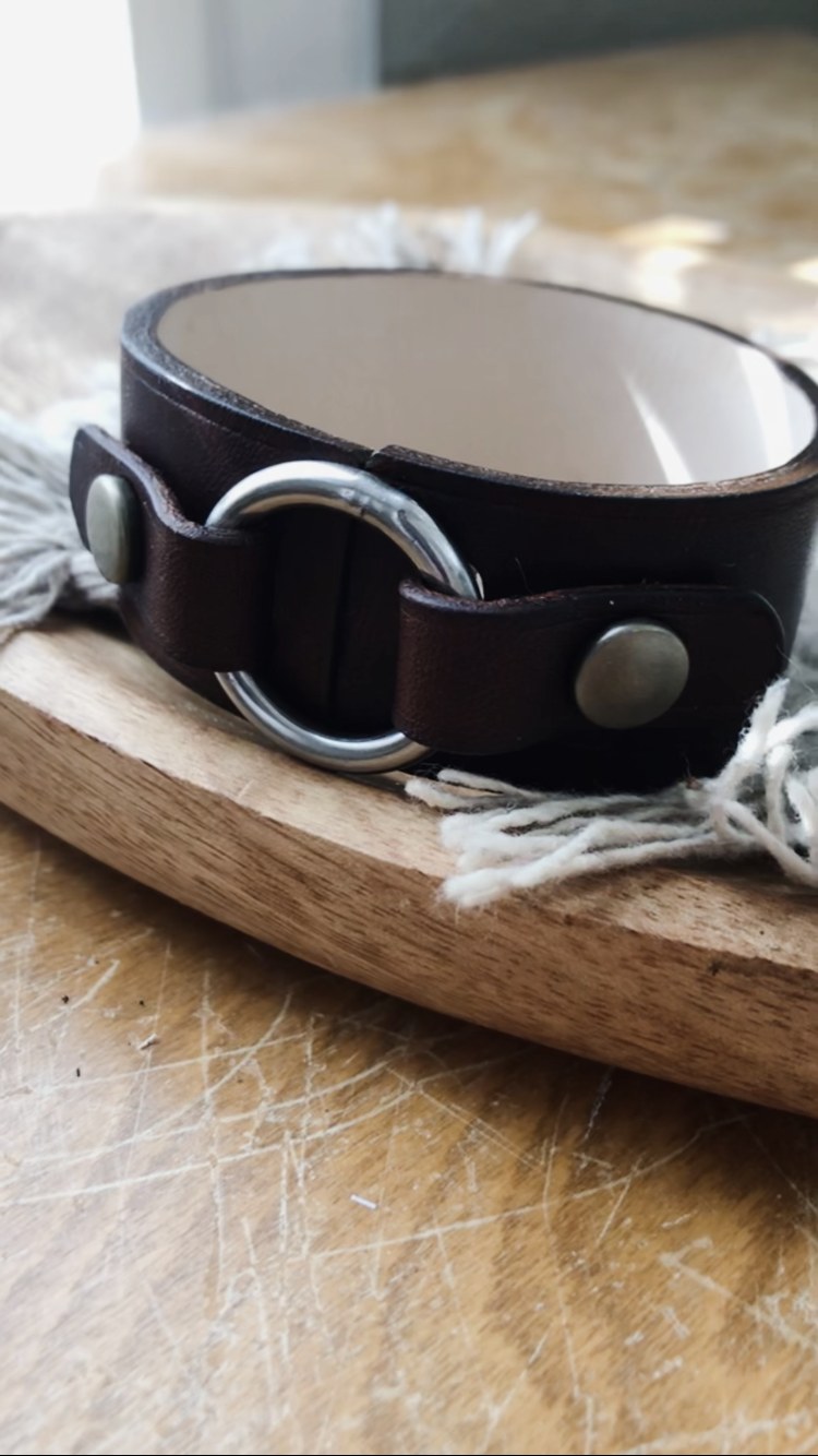 Handcrafted dark brown leather bracelet with silver metal ring and snap closure, resting on a wooden surface with natural light highlighting the texture
